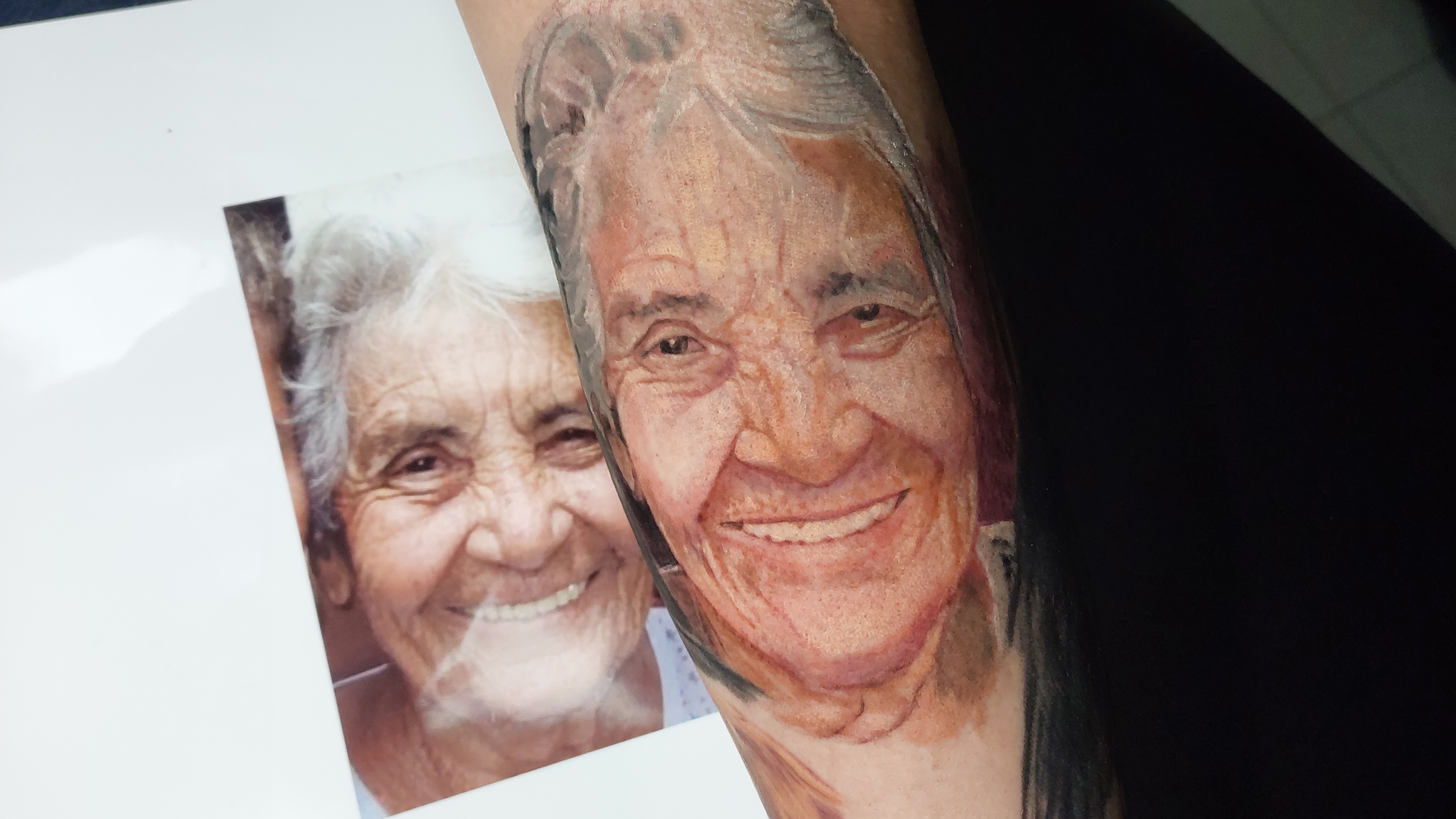 Retrato realista colorido de senhora com foto de referência ao lado, trabalho autoral do tatuador
